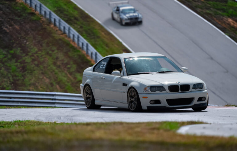 Closeup of a white sedan driving on the Ozark International HPDE track, with another vehicle coming down a steep hill in the background.